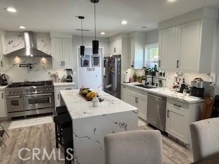 kitchen with wall chimney exhaust hood, hanging light fixtures, a kitchen island, stainless steel appliances, and white cabinets