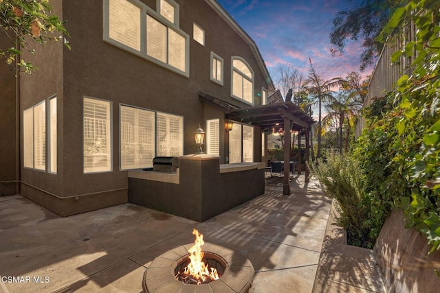 property exterior at dusk featuring an outdoor kitchen, a pergola, a patio area, and a fire pit