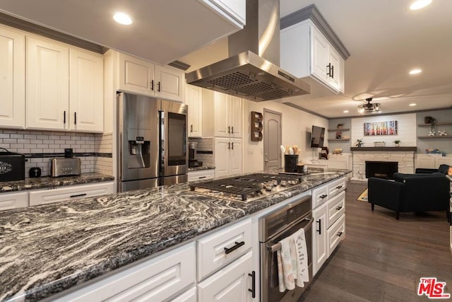 kitchen with appliances with stainless steel finishes, white cabinetry, island range hood, a stone fireplace, and dark stone counters