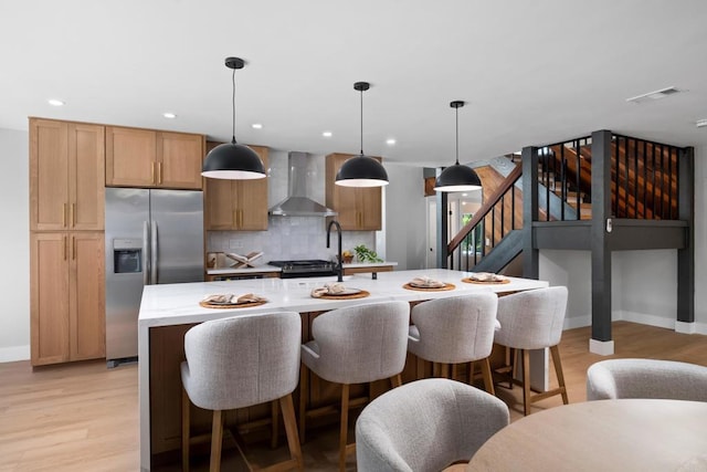 kitchen featuring pendant lighting, a kitchen island with sink, light hardwood / wood-style floors, stainless steel fridge with ice dispenser, and wall chimney exhaust hood