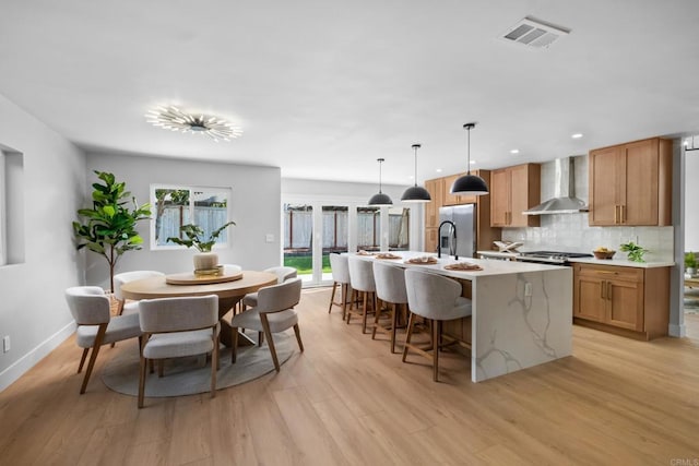 kitchen featuring wall chimney exhaust hood, a center island with sink, light wood-type flooring, pendant lighting, and backsplash