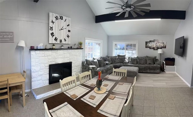 tiled dining space with ceiling fan, a stone fireplace, high vaulted ceiling, and beamed ceiling