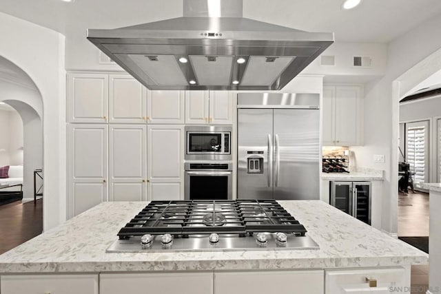 kitchen with built in appliances, light stone counters, white cabinets, and island exhaust hood