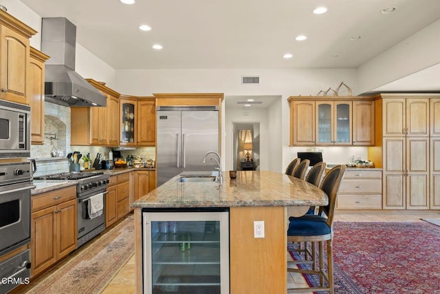 kitchen with beverage cooler, a kitchen breakfast bar, built in appliances, a center island with sink, and wall chimney range hood