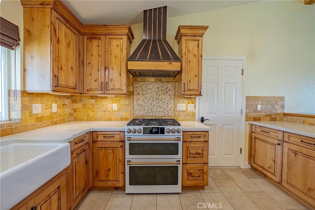 kitchen featuring wall chimney exhaust hood, tasteful backsplash, light countertops, brown cabinets, and range with two ovens