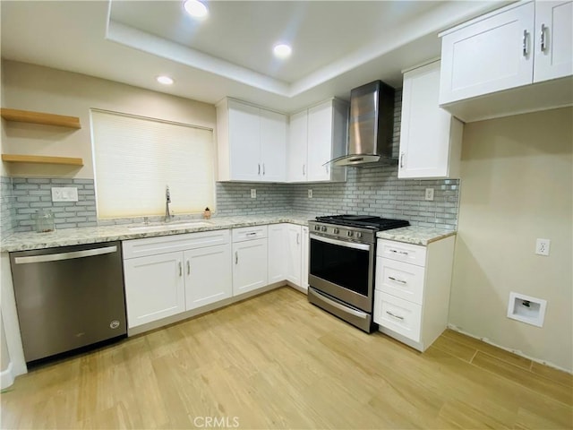 kitchen with wall chimney range hood, sink, stainless steel appliances, a tray ceiling, and white cabinets