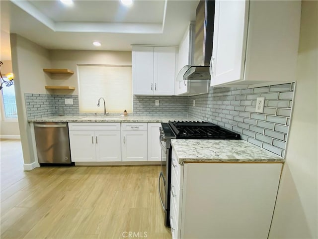 kitchen featuring white cabinetry, a tray ceiling, stainless steel appliances, wall chimney range hood, and light hardwood / wood-style flooring