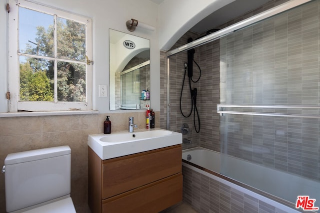 full bathroom with bath / shower combo with glass door, tile walls, vanity, and toilet