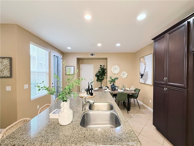 kitchen with an island with sink, sink, light tile patterned floors, light stone counters, and dark brown cabinets