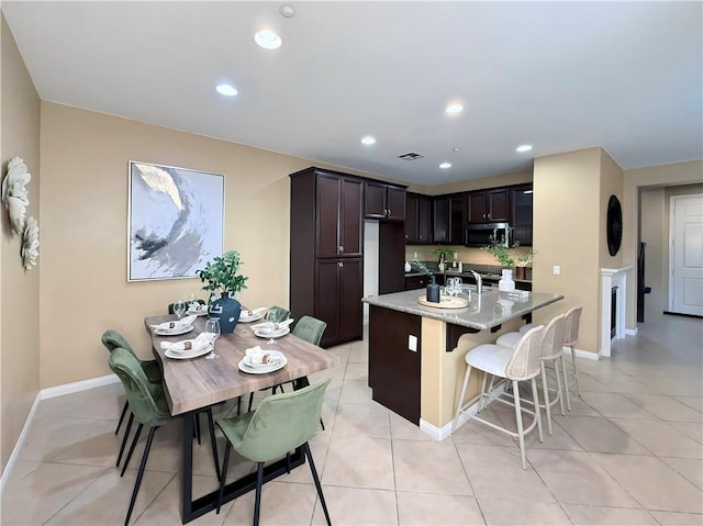 kitchen with light tile patterned floors, sink, a kitchen island with sink, light stone counters, and dark brown cabinetry