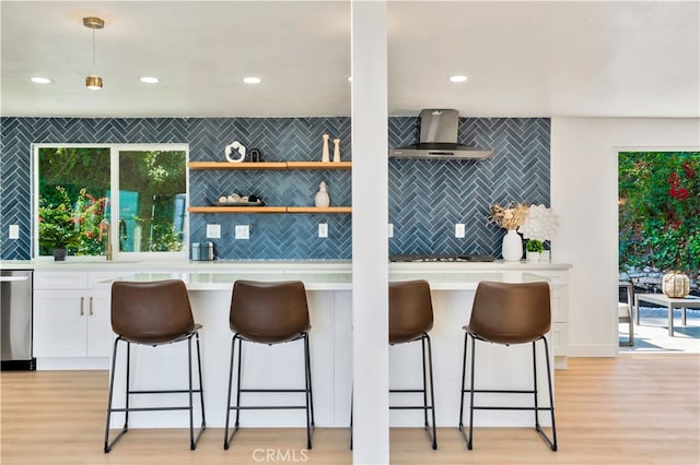 kitchen featuring a breakfast bar area, dishwasher, hanging light fixtures, white cabinets, and wall chimney exhaust hood