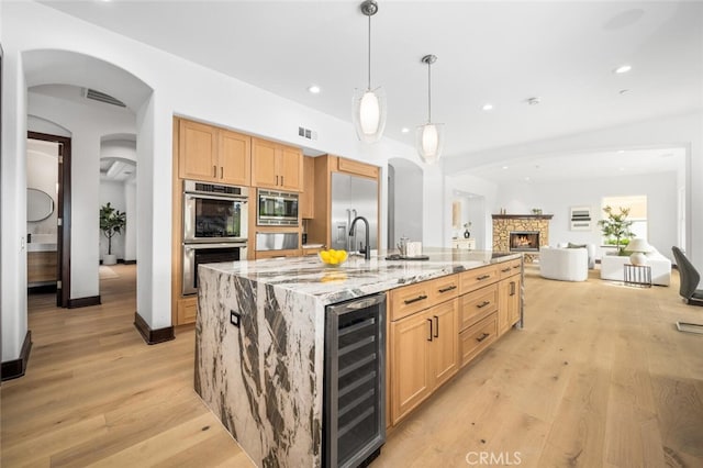 kitchen with decorative light fixtures, wine cooler, a kitchen island with sink, built in appliances, and light stone countertops