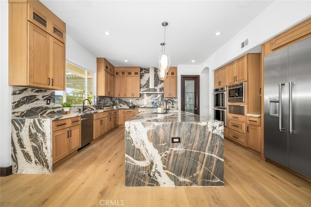 kitchen featuring a spacious island, wall chimney exhaust hood, built in appliances, pendant lighting, and light stone countertops