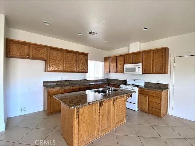 kitchen with an island with sink, sink, dark stone countertops, light tile patterned floors, and white appliances