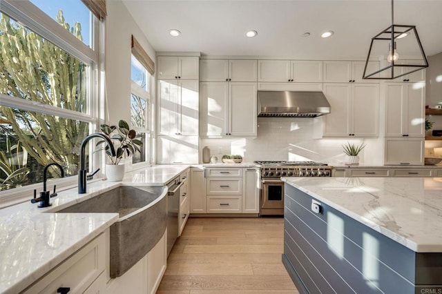kitchen with light stone counters, a sink, light wood-type flooring, wall chimney exhaust hood, and appliances with stainless steel finishes