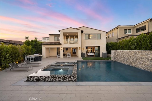 back house at dusk with a balcony, a pool with hot tub, and a patio area