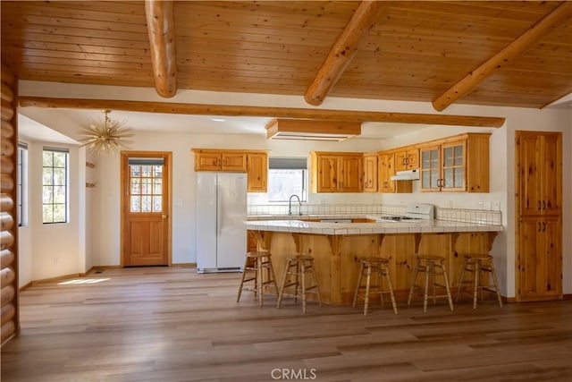 kitchen with tile countertops, freestanding refrigerator, under cabinet range hood, a kitchen bar, and a sink