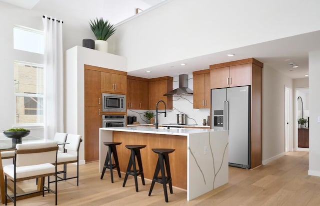 kitchen with light wood-type flooring, a kitchen breakfast bar, stainless steel appliances, a kitchen island with sink, and wall chimney range hood