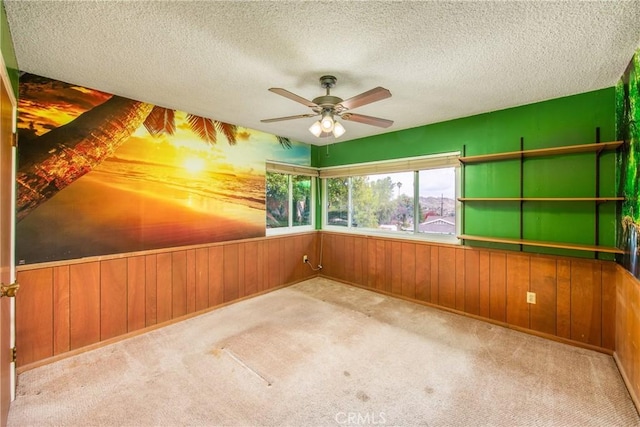 carpeted empty room with a textured ceiling, a ceiling fan, wood walls, and wainscoting