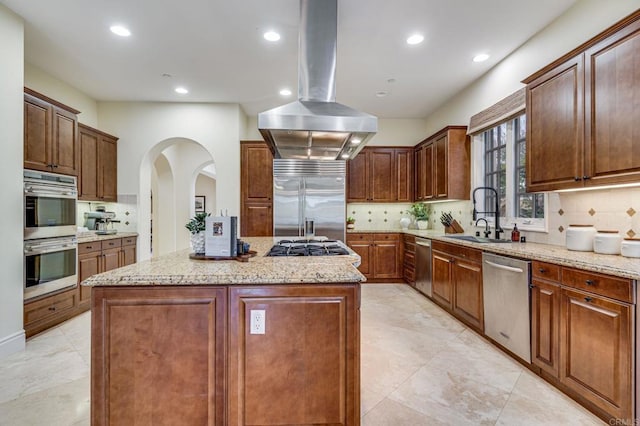 kitchen featuring stainless steel appliances, light stone counters, island range hood, a kitchen island, and sink