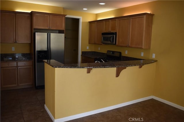 kitchen with a breakfast bar area, appliances with stainless steel finishes, dark tile patterned flooring, kitchen peninsula, and dark stone counters