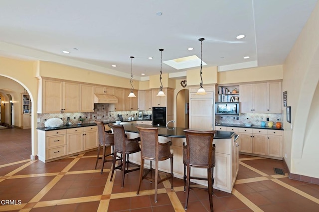 kitchen featuring arched walkways, a breakfast bar area, dark countertops, light brown cabinets, and under cabinet range hood