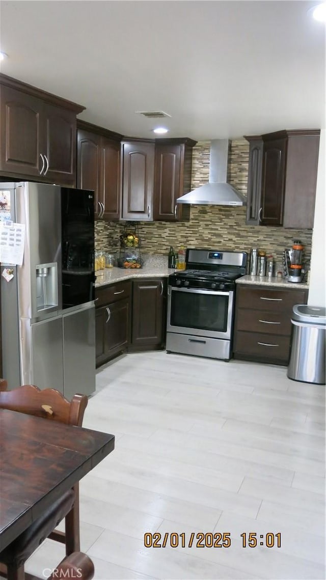 kitchen featuring decorative backsplash, stainless steel appliances, dark brown cabinets, wall chimney range hood, and light wood-type flooring