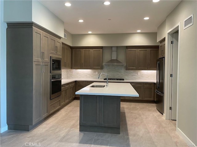 kitchen featuring wall chimney range hood, sink, a kitchen island with sink, stainless steel appliances, and tasteful backsplash