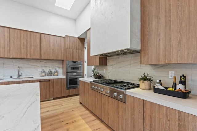 kitchen with appliances with stainless steel finishes, a skylight, sink, wall chimney range hood, and light hardwood / wood-style flooring