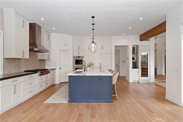 kitchen featuring wall chimney range hood, white cabinetry, hanging light fixtures, stainless steel appliances, and a center island with sink