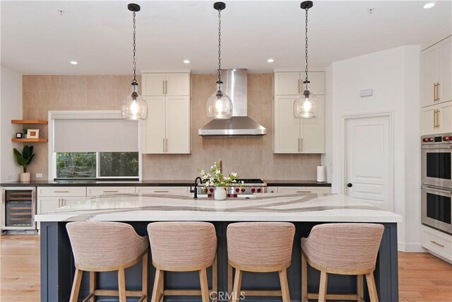 kitchen with light wood finished floors, beverage cooler, stainless steel double oven, white cabinets, and wall chimney exhaust hood