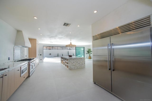 kitchen with custom range hood, built in appliances, light brown cabinetry, and light stone counters