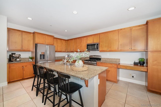 kitchen with a kitchen bar, light stone counters, built in desk, stainless steel appliances, and a kitchen island with sink