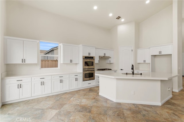 kitchen with a towering ceiling, an island with sink, sink, white cabinets, and stainless steel double oven