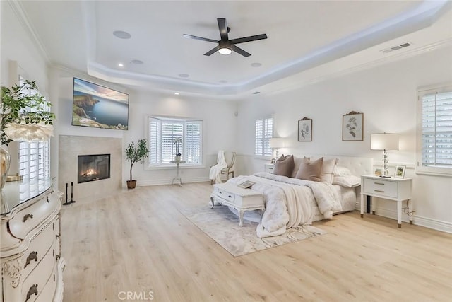 bedroom with crown molding, ceiling fan, a tray ceiling, a premium fireplace, and light hardwood / wood-style flooring