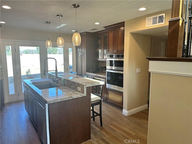 kitchen with sink, light stone counters, a center island with sink, decorative light fixtures, and stainless steel double oven