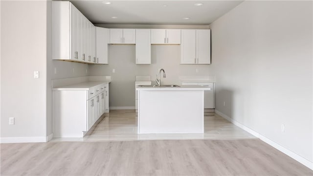 kitchen featuring baseboards, a center island with sink, light wood finished floors, a sink, and white cabinets