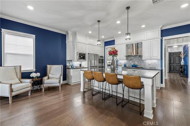 kitchen with pendant lighting, a kitchen island with sink, stainless steel appliances, white cabinets, and wall chimney exhaust hood