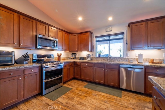 kitchen with sink, vaulted ceiling, light wood-type flooring, stainless steel appliances, and light stone countertops