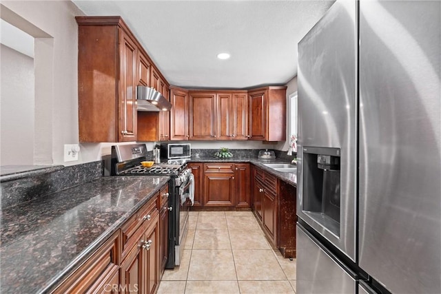 kitchen featuring light tile patterned floors, dark stone counters, appliances with stainless steel finishes, wall chimney range hood, and a sink