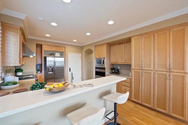 kitchen with crown molding, a breakfast bar, appliances with stainless steel finishes, wall chimney exhaust hood, and light wood-type flooring