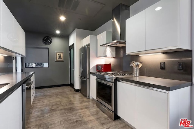 kitchen featuring white cabinetry, decorative backsplash, stainless steel appliances, light wood-type flooring, and wall chimney exhaust hood