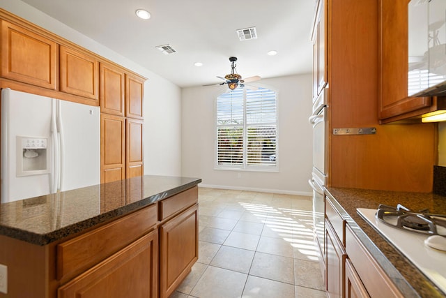 kitchen featuring light tile patterned flooring, ceiling fan, a kitchen island, white appliances, and dark stone counters