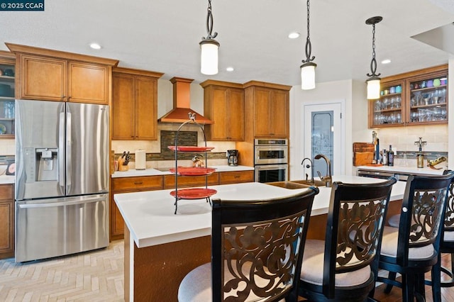 kitchen with pendant lighting, stainless steel appliances, an island with sink, and custom range hood