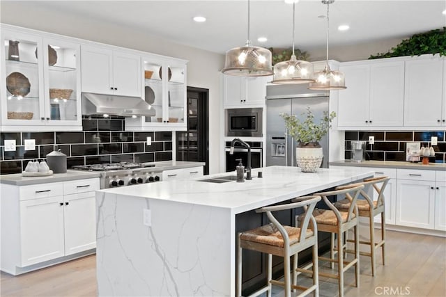 kitchen featuring a kitchen island with sink, built in appliances, white cabinetry, and ventilation hood