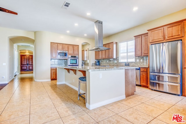 kitchen with a breakfast bar area, appliances with stainless steel finishes, island exhaust hood, light stone counters, and a kitchen island