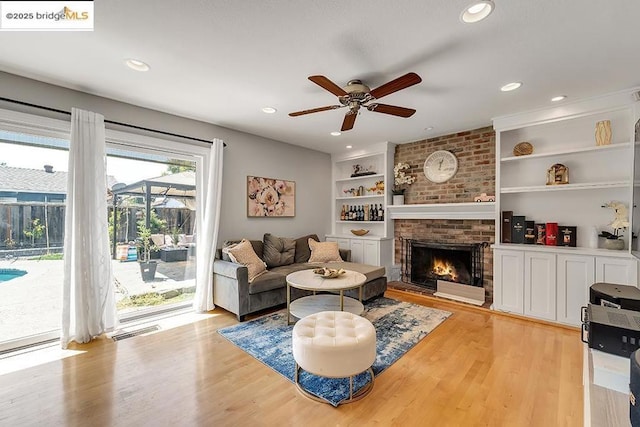 living room with ceiling fan, a brick fireplace, built in features, and light wood-type flooring