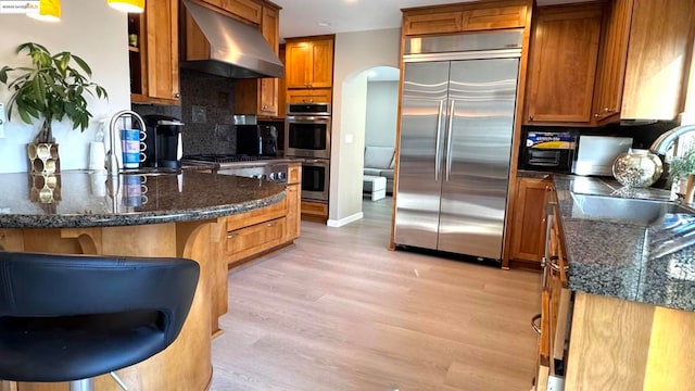 kitchen with sink, a kitchen breakfast bar, stainless steel appliances, dark stone counters, and wall chimney exhaust hood