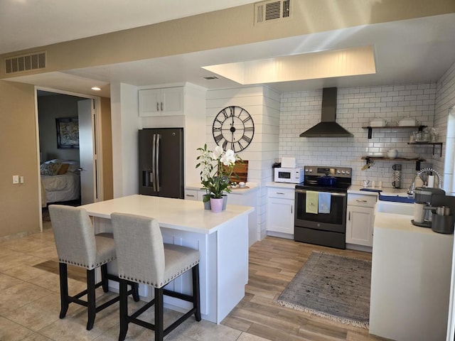 kitchen featuring wall chimney exhaust hood, a breakfast bar area, black fridge, electric stove, and white cabinets