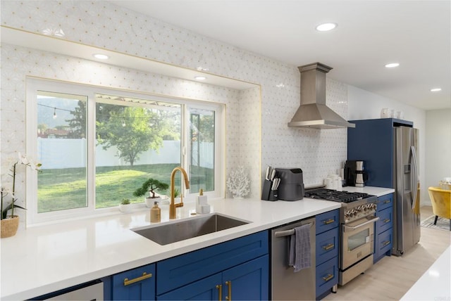 kitchen featuring appliances with stainless steel finishes, blue cabinets, sink, and wall chimney range hood